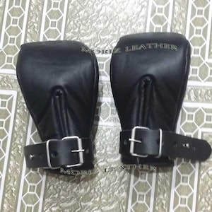 Può includere: Un paio di manette in pelle nera con fibbie argentate. Le manette hanno una parte superiore arrotondata e un cinturino che si allaccia al polso. Il testo "MORIZ LEATHER" è stampato sulle manette.