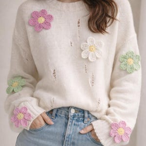 Può includere: Maglione color crema con dettagli strappati e applicazioni floreali all'uncinetto in rosa, bianco e verde. I fiori hanno centri gialli. Il maglione è abbinato a jeans di denim blu chiaro.