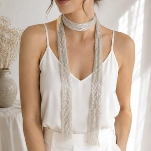 May include: A silver scarf with a delicate, shimmering design. The scarf is draped around the neck and hangs down the front. The scarf is made of a lightweight material and has a subtle sheen. The scarf is a stylish accessory.