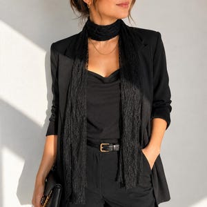 May include: A woman in an all-black ensemble: blazer, top, trousers, and a lace scarf. The outfit is accessorized with a black belt and a gold necklace. A black clutch is held in the left hand.
