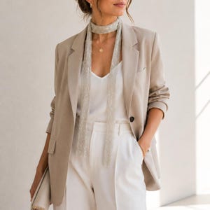 May include: A woman's outfit featuring a beige blazer, white top, and white pants. A silver beaded scarf is draped around the neck. The outfit is accessorized with a gold necklace and bracelet. A beige clutch is held in the left hand.