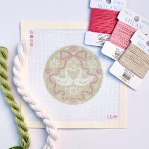 Puede incluir: Un kit de punto de aguja con un diseño circular con dos cisnes blancos, lazos rosas y corazones sobre un fondo verde claro. Incluye carretes de hilo rosa y beige, e hilo de bordar verde y blanco.