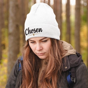 May include: A white knitted beanie featuring the word "Chosen" in black script and a small cross design. The beanie is worn by a person with long brown hair, wearing a black jacket, set against a blurred forest backdrop.