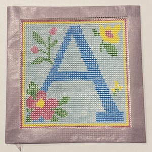 Peut inclure: Carré de point de croix avec la lettre "A" bleue et des motifs floraux. La lettre est centrée sur un fond bleu clair, entourée de fleurs roses, jaunes et vertes. Le motif est encadré par une bordure jaune et un tissu lavande.