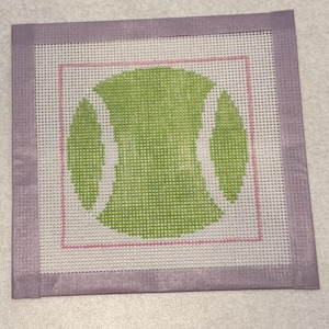 May include: A square needlepoint canvas with a tennis ball design. The tennis ball is green with white accents, set within a pink and lavender border. The canvas is made of a white mesh material.