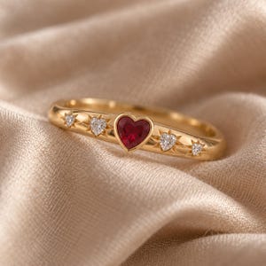 Può includere: Un anello in oro con una pietra preziosa rossa centrale a forma di cuore, affiancata da due pietre chiare più piccole a forma di cuore e due minuscole pietre rotonde chiare, incastonate in incisioni a stella sulla superficie dorata lucida.