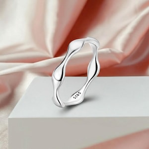 May include: A silver ring with a flowing, organic design. The ring has a wavy, fluid shape, with a polished finish. The inside of the ring is stamped with "S925". The ring is displayed on a white surface with a pink background.