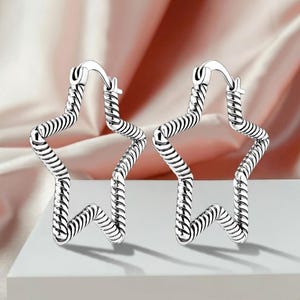 Puede incluir: Un par de pendientes de aro plateados con forma de estrella. Los pendientes tienen un diseño texturizado en espiral y un cierre de bisagra. El fondo es de tela rosa suave.