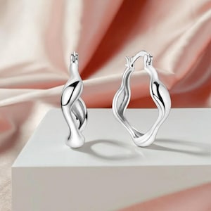 May include: A pair of silver hoop earrings with a distinctive, flowing design. The earrings feature a twisted, organic shape, with a smooth, polished finish. The design is modern and elegant, suitable for everyday wear or special occasions.