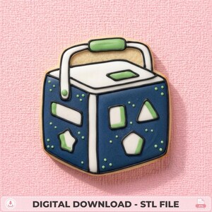 May include: A cookie shaped like a blue and white toy shape sorter cube with a green handle. The cube features various cutouts including a star, triangle, rectangle, and pentagon, outlined in black icing with small green dots on the blue surfaces.