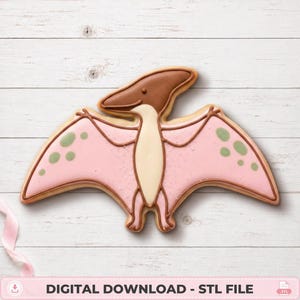 Peut inclure: Un biscuit en forme de ptérodactyle décoré avec un glaçage rose clair sur les ailes, agrémenté de petites taches vertes. Le corps est de couleur crème et la tête est brune. Le biscuit présente des contours en glaçage brun et une texture.