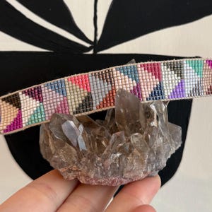 May include: A colorful beaded bracelet with a geometric pattern in shades of pink, purple, blue, and beige. The bracelet is displayed on a cluster of crystals. The bracelet is approximately 0.75 inches wide.