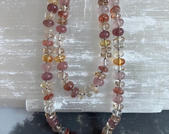 May include: A multi-colored gemstone necklace with round beads in shades of pink, orange, and clear. The necklace is made of natural stones and is strung on a silver chain.