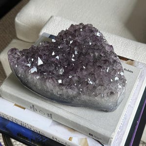 May include: A heart-shaped amethyst crystal cluster featuring deep purple, faceted points that transition into a translucent, pale base. The mineral specimen displays a natural, jagged texture with varying shades of violet throughout the crystalline structure.