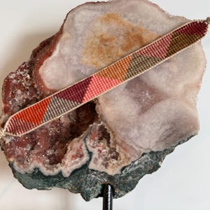 May include: A handmade friendship bracelet with a woven pattern of orange, red, pink, and olive green. The bracelet is displayed on a large, multi-colored geode. The bracelet is approximately 0.5 inches wide.