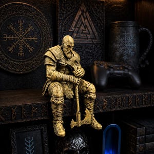 Peut inclure: Une figurine dorée représentant un guerrier barbu assis tenant une hache, placée devant une étagère sombre ornée d'un emblème circulaire Vegvisir, d'une plaque triangulaire Valknut, d'une tasse noire à motif flocon et d'une sculpture de crâne.