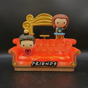 May include: A miniature orange couch with the word "FRIENDS" on the front, featuring two small figurines. The couch has a gold-colored back and is set against a dark background. The figurines are cartoon-like characters.