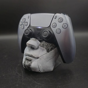 Könnte beinhalten: Ein grauer PlayStation 5-Controller liegt auf einem grau-weißen, geformten Kopf. Der Controller hat schwarze Tasten und Richtungspads. Der Kopf hat ein strukturiertes, verwittertes Aussehen mit dunklen Akzenten.
