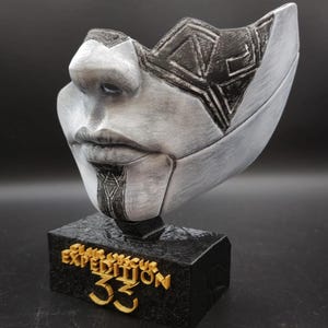 Op de afbeelding: Een zilver-zwart maskerbeeld met een gestileerd gezicht, geplaatst op een zwarte basis. De basis heeft gouden tekst met de tekst "Expedition 33". Het masker heeft geometrische patronen en een gedetailleerde gezichtsstructuur.