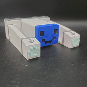 以下が含まれることがあります： ブロックデザインの白と青のおもちゃのフィギュア。フィギュアは、黒い正方形の目と笑顔の口を持つ青い顔をしています。フィギュアには、緑色の足跡が付いた長方形の腕と足があります。おもちゃは暗い表面にあります。