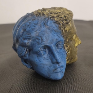 Pode incluir: Uma escultura de face dupla apresentando duas cabe&ccedil;as humanas unidas costas com costas. Um lado tem acabamento em azul fosco com tra&ccedil;os faciais e cabelo detalhados, enquanto o outro lado tem acabamento em dourado met&aacute;lico com superf&iacute;cie texturizada.