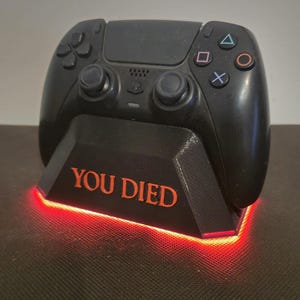 May include: Black PlayStation 5 controller resting on a black stand with the words "YOU DIED" in orange. The stand has a red glowing light around the base. Gaming accessory.