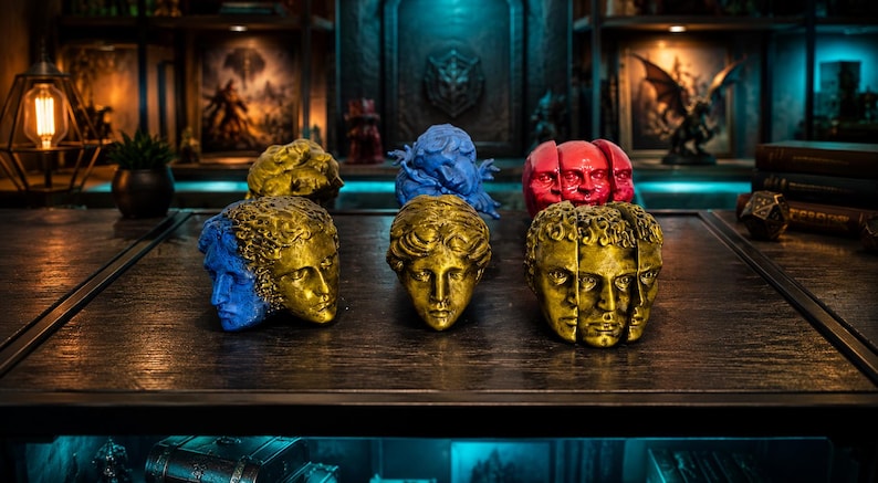 Pode incluir: Uma cole&ccedil;&atilde;o de ornamentos de cabe&ccedil;a esculturais com m&uacute;ltiplas faces e designs divididos em acabamentos de ouro met&aacute;lico, azul vibrante e vermelho brilhante. Estes objetos decorativos exibem tra&ccedil;os faciais intrincados e cabelo texturizado.