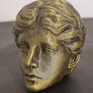 Pode incluir: Um busto escultural de uma cabe&ccedil;a humana com acabamento em tom dourado met&aacute;lico e sombreamento antigo escuro. A figura apresenta cabelo ondulado penteado para tr&aacute;s, tra&ccedil;os faciais definidos incluindo um nariz reto e uma express&atilde;o neutra.