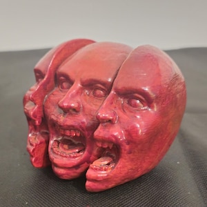 Pode incluir: Uma escultura vermelha apresentando tr&ecirc;s cabe&ccedil;as humanas interconectadas dispostas em fila. Cada cabe&ccedil;a exibe uma express&atilde;o facial distinta com boca aberta, dentes vis&iacute;veis e olhos detalhados. A superf&iacute;cie tem uma textura.