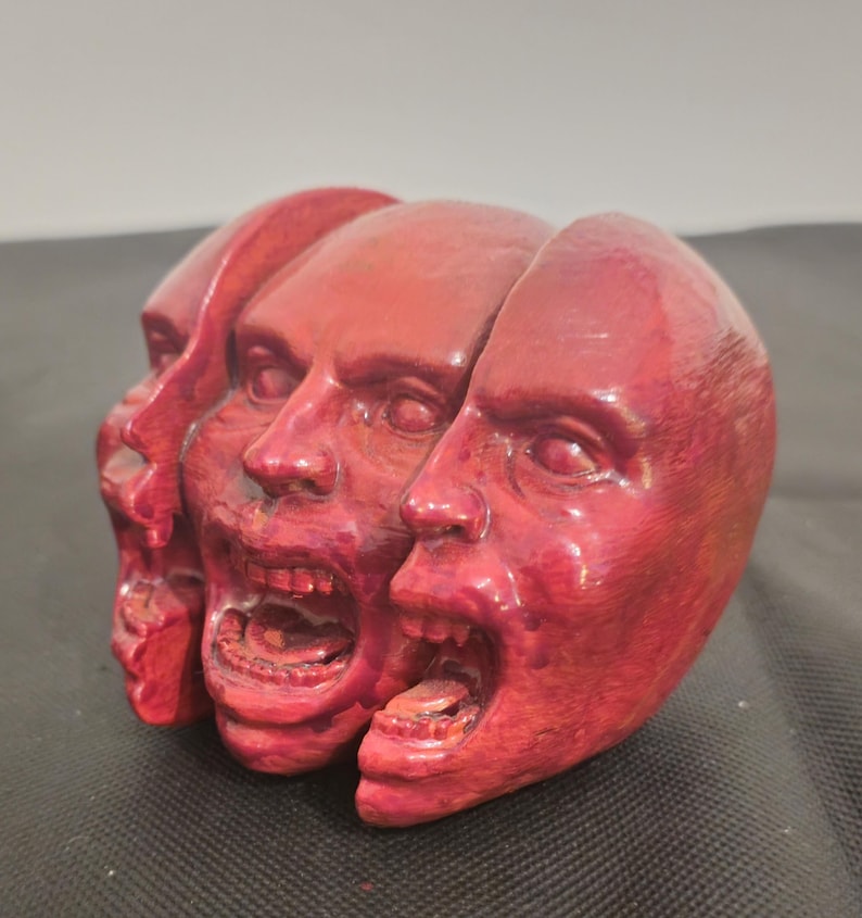 Pode incluir: Uma escultura vermelha apresentando tr&ecirc;s cabe&ccedil;as humanas interconectadas dispostas em fila. Cada cabe&ccedil;a exibe uma express&atilde;o facial distinta com boca aberta, dentes vis&iacute;veis e olhos detalhados. A superf&iacute;cie tem uma textura.
