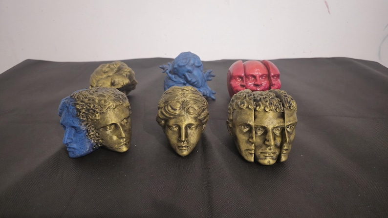 Pode incluir: Uma cole&ccedil;&atilde;o de seis pequenas cabe&ccedil;as esculturais em v&aacute;rias cores e acabamentos. O grupo inclui pe&ccedil;as douradas, azuis e vermelhas, algumas com rostos m&uacute;ltiplos ou designs divididos, exibindo tra&ccedil;os faciais detalhados e texturas.