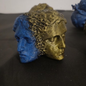 Pode incluir: Uma escultura de face dupla apresentando duas cabe&ccedil;as humanas unidas costas com costas. Um lado tem acabamento em azul fosco, enquanto o outro lado tem acabamento em ouro met&aacute;lico. O cabelo em ambas as cabe&ccedil;as tem uma apar&ecirc;ncia texturizada.