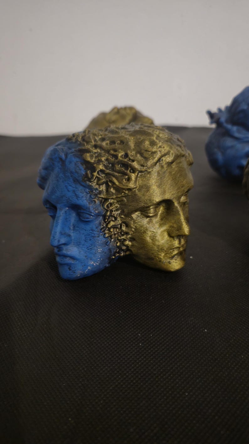 Pode incluir: Uma escultura de face dupla apresentando duas cabe&ccedil;as humanas unidas costas com costas. Um lado tem acabamento em azul fosco, enquanto o outro lado tem acabamento em ouro met&aacute;lico. O cabelo em ambas as cabe&ccedil;as tem uma apar&ecirc;ncia texturizada.