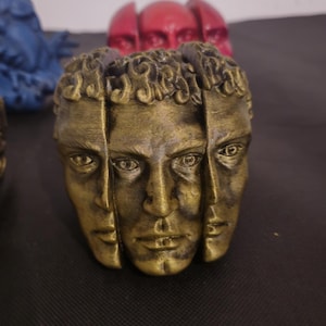 Pode incluir: Uma escultura de cor dourada apresentando tr&ecirc;s rostos humanos distintos dispostos em um padr&atilde;o circular. Cada rosto exibe tra&ccedil;os detalhados e cabelos cacheados, com um acabamento met&aacute;lico desgastado que destaca o contorno dos olhos e l&aacute;bios.