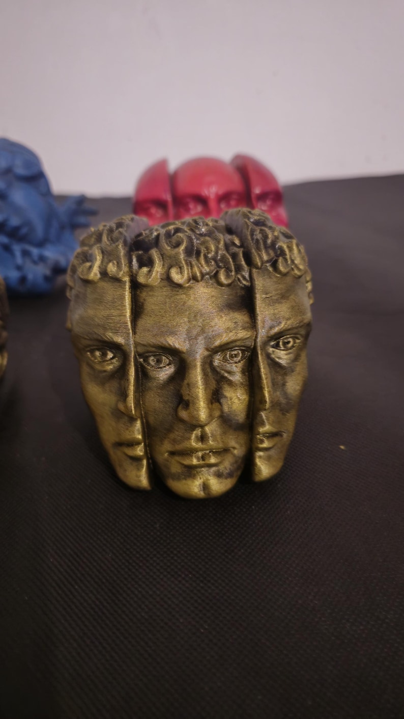 Pode incluir: Uma escultura de cor dourada apresentando tr&ecirc;s rostos humanos distintos dispostos em um padr&atilde;o circular. Cada rosto exibe tra&ccedil;os detalhados e cabelos cacheados, com um acabamento met&aacute;lico desgastado que destaca o contorno dos olhos e l&aacute;bios.