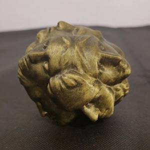 Pode incluir: Uma escultura esf&eacute;rica com acabamento em bronze met&aacute;lico, apresentando m&uacute;ltiplos rostos humanos integrados na sua superf&iacute;cie. O objeto exibe uma apar&ecirc;ncia texturizada e desgastada com reflexos escuros nas cavidades e tons claros nos relevos.