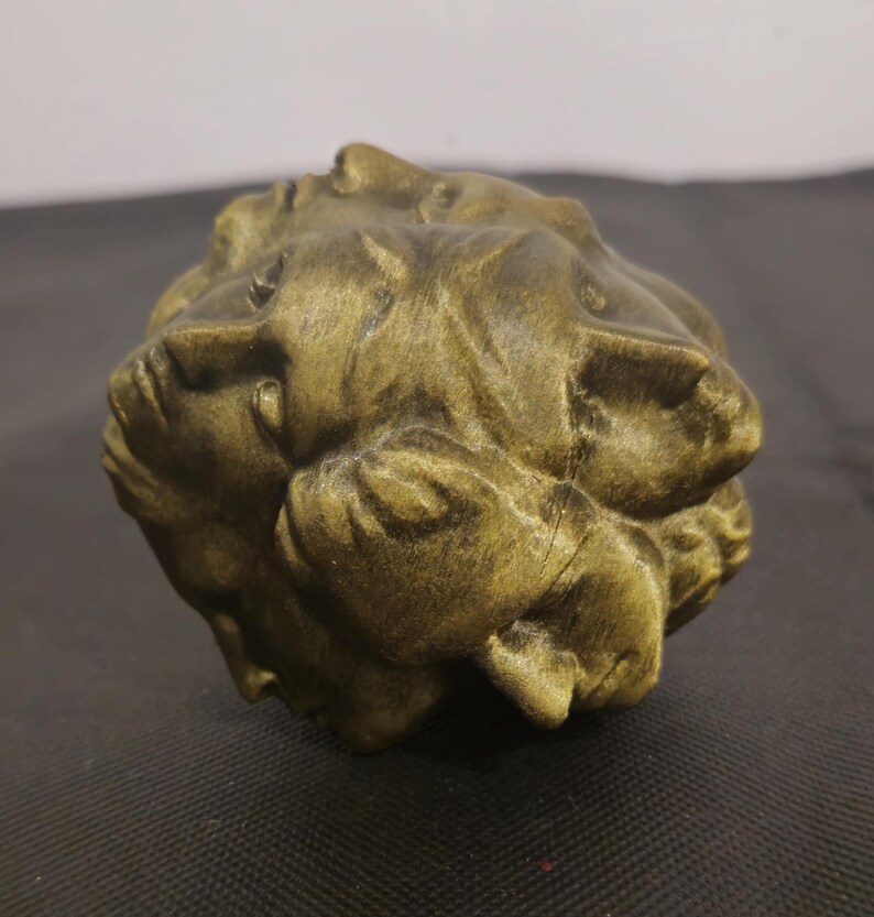 Pode incluir: Uma escultura esf&eacute;rica com acabamento em bronze met&aacute;lico, apresentando m&uacute;ltiplos rostos humanos integrados na sua superf&iacute;cie. O objeto exibe uma apar&ecirc;ncia texturizada e desgastada com reflexos escuros nas cavidades e tons claros nos relevos.