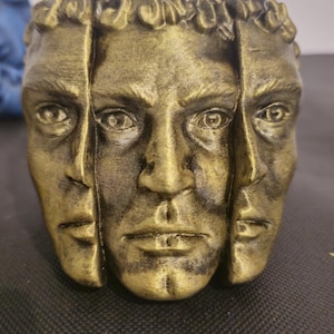Pode incluir: Um vaso cil&iacute;ndrico com acabamento dourado met&aacute;lico apresentando tr&ecirc;s rostos humanos esculpidos dispostos ao redor do exterior. Os rostos s&atilde;o detalhados com olhos, narizes e l&aacute;bios definidos, e a borda superior mostra padr&otilde;es de cabelo texturizados.