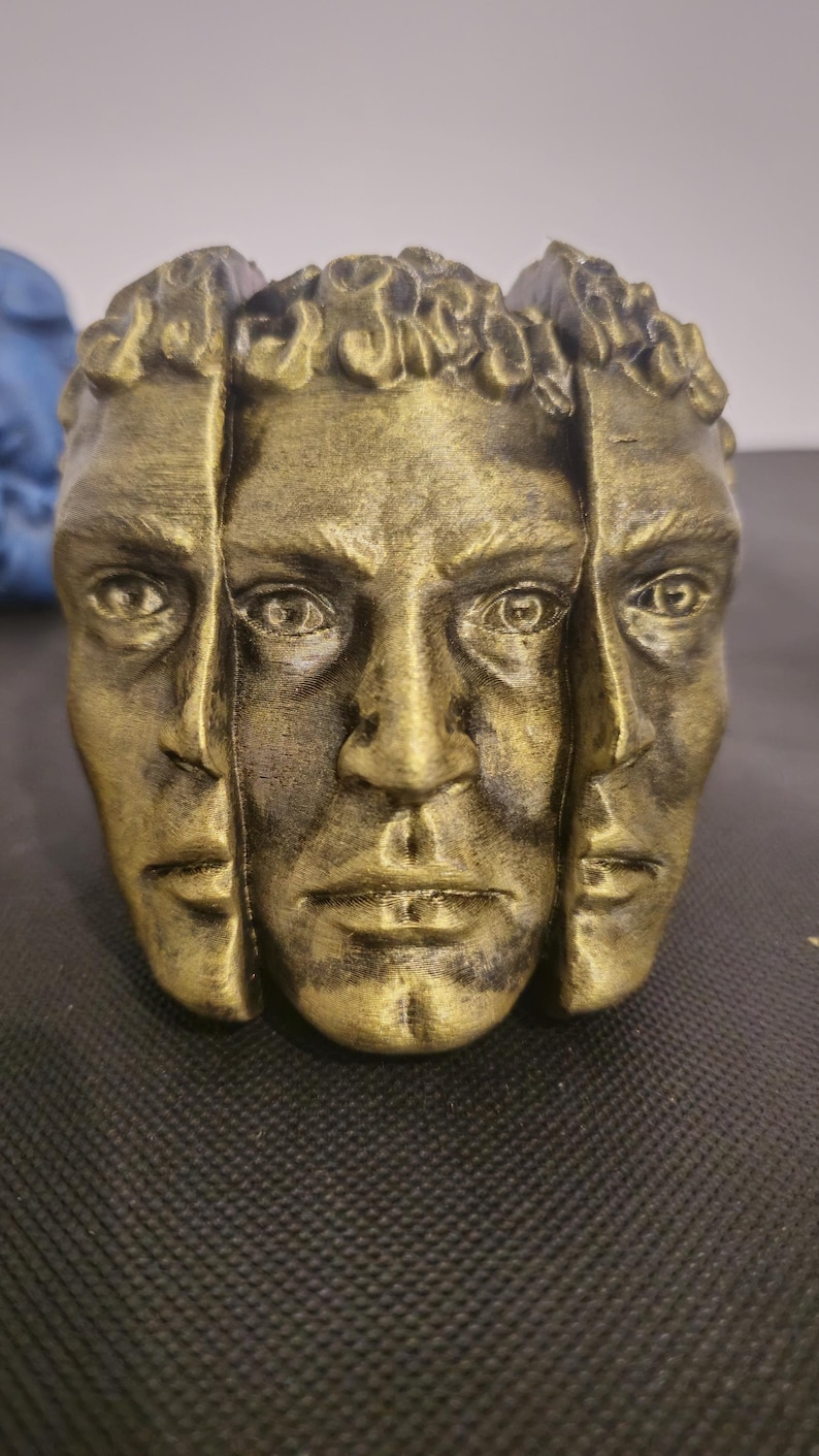 Pode incluir: Um vaso cil&iacute;ndrico com acabamento dourado met&aacute;lico apresentando tr&ecirc;s rostos humanos esculpidos dispostos ao redor do exterior. Os rostos s&atilde;o detalhados com olhos, narizes e l&aacute;bios definidos, e a borda superior mostra padr&otilde;es de cabelo texturizados.