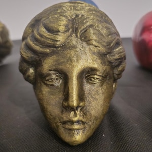 Pode incluir: Uma escultura de cor bronze de uma cabe&ccedil;a humana com tra&ccedil;os faciais detalhados e cabelo ondulado. A superf&iacute;cie tem um acabamento met&aacute;lico texturizado com destaques escuros nas fendas, criando uma apar&ecirc;ncia de desgaste natural.