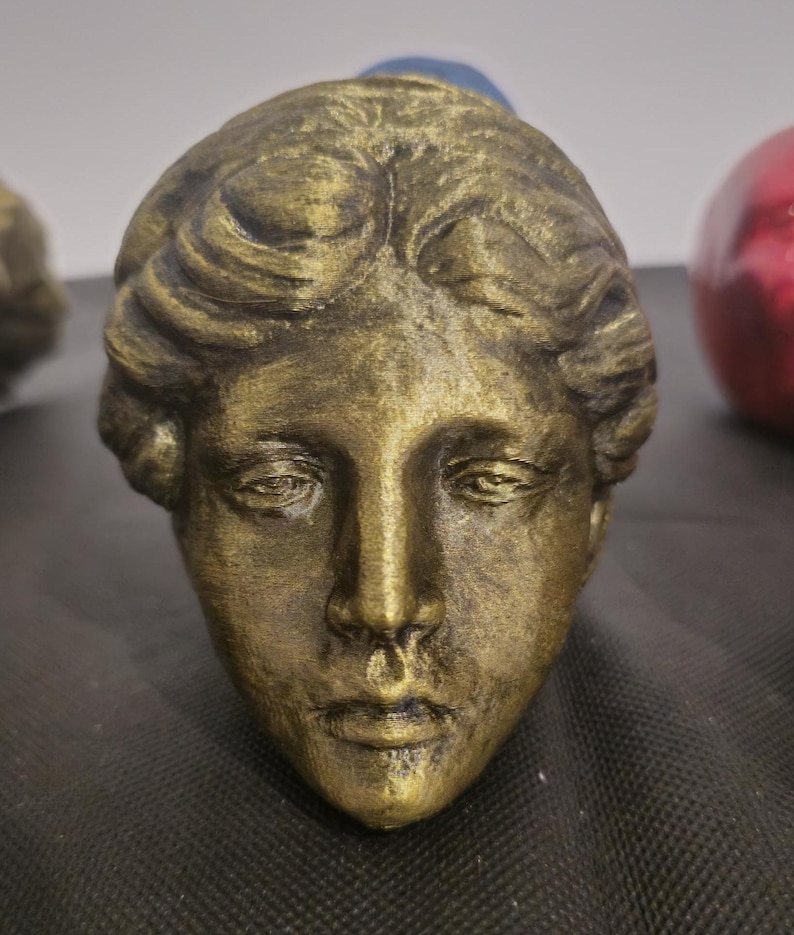 Pode incluir: Uma escultura de cor bronze de uma cabe&ccedil;a humana com tra&ccedil;os faciais detalhados e cabelo ondulado. A superf&iacute;cie tem um acabamento met&aacute;lico texturizado com destaques escuros nas fendas, criando uma apar&ecirc;ncia de desgaste natural.