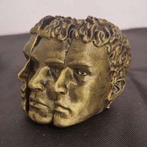 Pode incluir: Uma escultura decorativa apresentando tr&ecirc;s rostos humanos interconectados com um acabamento em ouro met&aacute;lico e reflexos antigos escuros. Os rostos est&atilde;o dispostos em um design de perfil sobreposto com tra&ccedil;os faciais detalhados e cabelo texturizado.