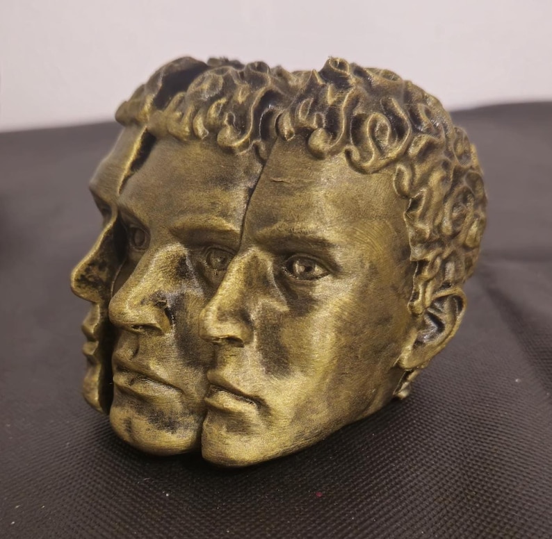 Pode incluir: Uma escultura decorativa apresentando tr&ecirc;s rostos humanos interconectados com um acabamento em ouro met&aacute;lico e reflexos antigos escuros. Os rostos est&atilde;o dispostos em um design de perfil sobreposto com tra&ccedil;os faciais detalhados e cabelo texturizado.