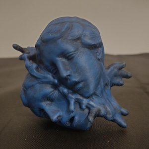 Pode incluir: Uma escultura azul mate representando dois rostos posicionados pr&oacute;ximos um do outro, com um rosto descansando sobre o outro. A superf&iacute;cie apresenta um acabamento texturizado e desgastado com sombras azul-escuras nos sulcos, criando profundidade.