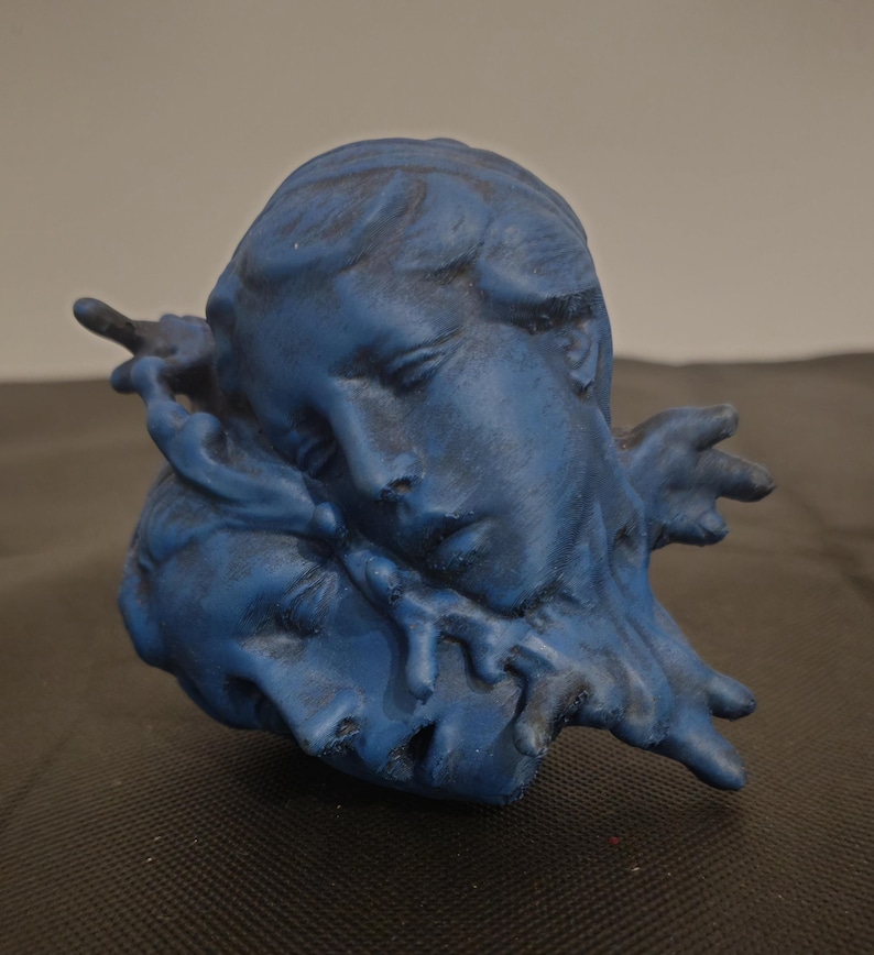 Pode incluir: Uma escultura azul mate representando dois rostos posicionados pr&oacute;ximos um do outro, com um rosto descansando sobre o outro. A superf&iacute;cie apresenta um acabamento texturizado e desgastado com sombras azul-escuras nos sulcos, criando profundidade.