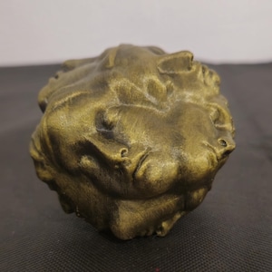 Pode incluir: Uma escultura esf&eacute;rica com acabamento em bronze met&aacute;lico, apresentando m&uacute;ltiplos rostos humanos moldados na superf&iacute;cie. Os rostos s&atilde;o retratados com olhos fechados e express&otilde;es neutras, criando um relevo texturizado e cont&iacute;nuo no objeto.
