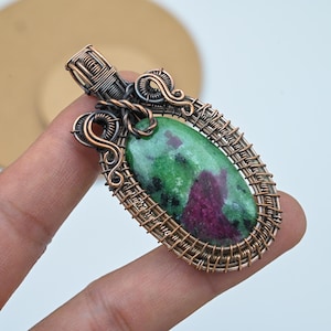 Könnte beinhalten: Ein handgefertigter Anhänger mit einem grünen und kastanienroten Zoisitstein, kunstvoll mit Kupfer- und Bronzetönen drahtgewickelt. Der oval geformte Stein ist der Mittelpunkt, wobei detaillierte Drahtarbeiten einen dekorativen Rahmen und eine Öse für die Halskette bilden.
