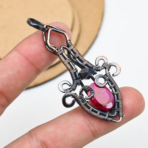 Könnte beinhalten: Ein handgefertigter Anhänger mit einem leuchtend roten Edelstein, der in einem detaillierten Drahtrahmen gefasst ist. Der Anhänger zeigt dunkle und kupferfarbene Metalle und schafft ein einzigartiges Design. Der Edelstein ist etwa 2 cm lang.