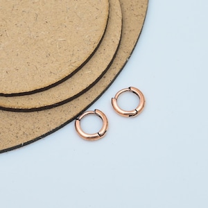 Könnte beinhalten: Zwei kleine, kupferfarbene Creolen. Die Ohrringe sind rund und haben ein schlichtes, klassisches Design. Sie werden auf einer weißen Oberfläche mit einem braunen, kreisförmigen Hintergrund präsentiert. Die Ohrringe haben eine warme, metallische Farbe.