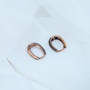Op de afbeelding: Een paar ovaalvormige oorringen met een koperkleurige afwerking. Elke oorbel heeft een getextureerde, kralenachtige buitenrand en een glad binnenoppervlak, ontworpen als een scharnierende ring die vastklikt voor veilig dragen aan de oorlel.